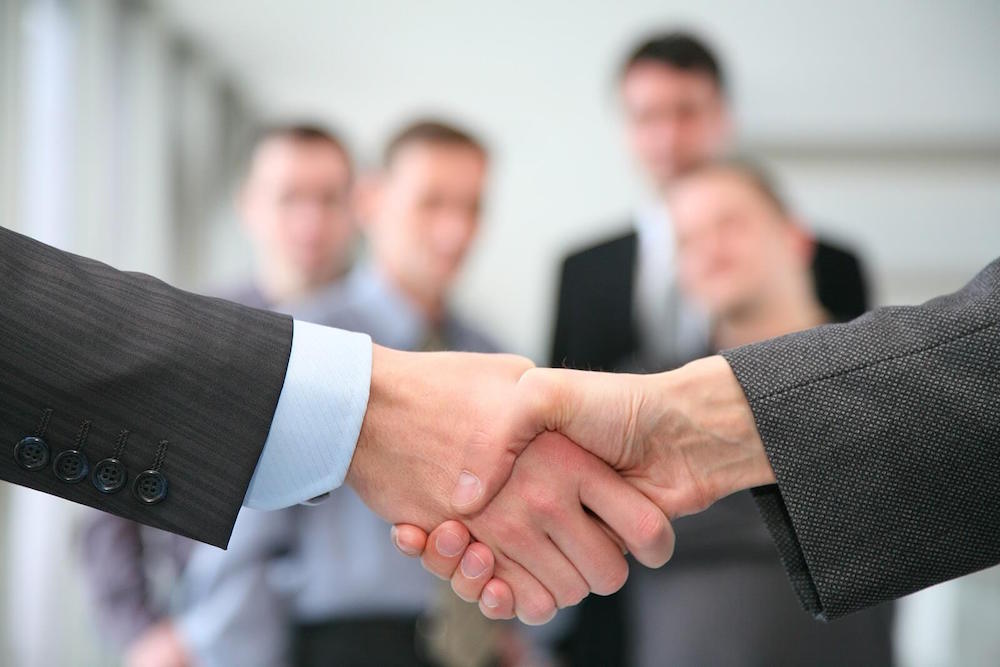 Two disembodied hands (man & woman’s) shaking hands with business professionals in the background